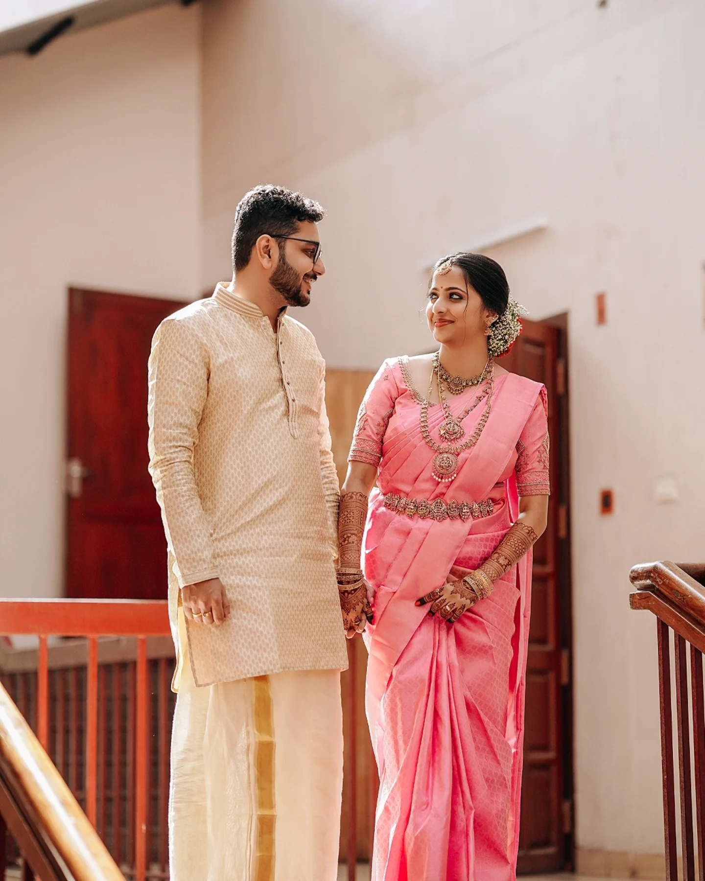 A smiling bride and groom in traditional wedding attire holding hands on a staircase