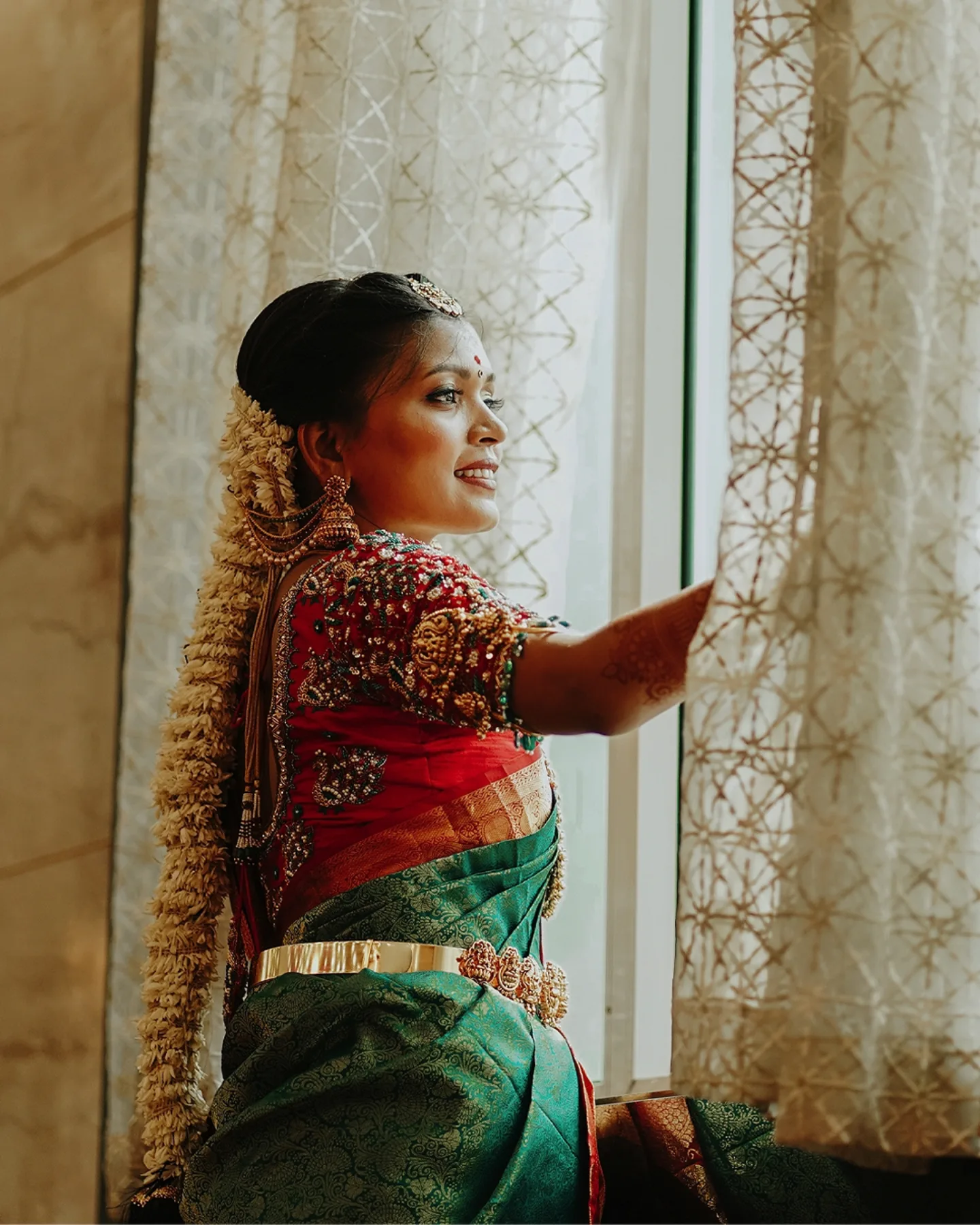 A bride in a green and red silk saree looking out a window